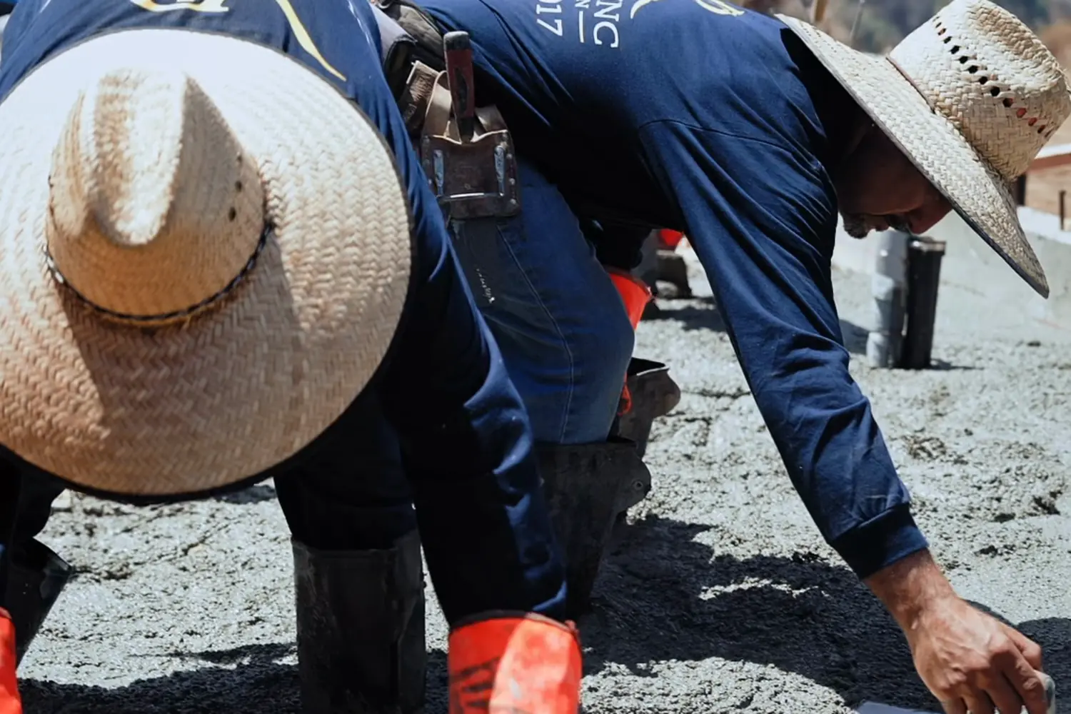 Two VR Plumbing Inc & Construction crew members in branded shirts and straw hats troweling wet concrete