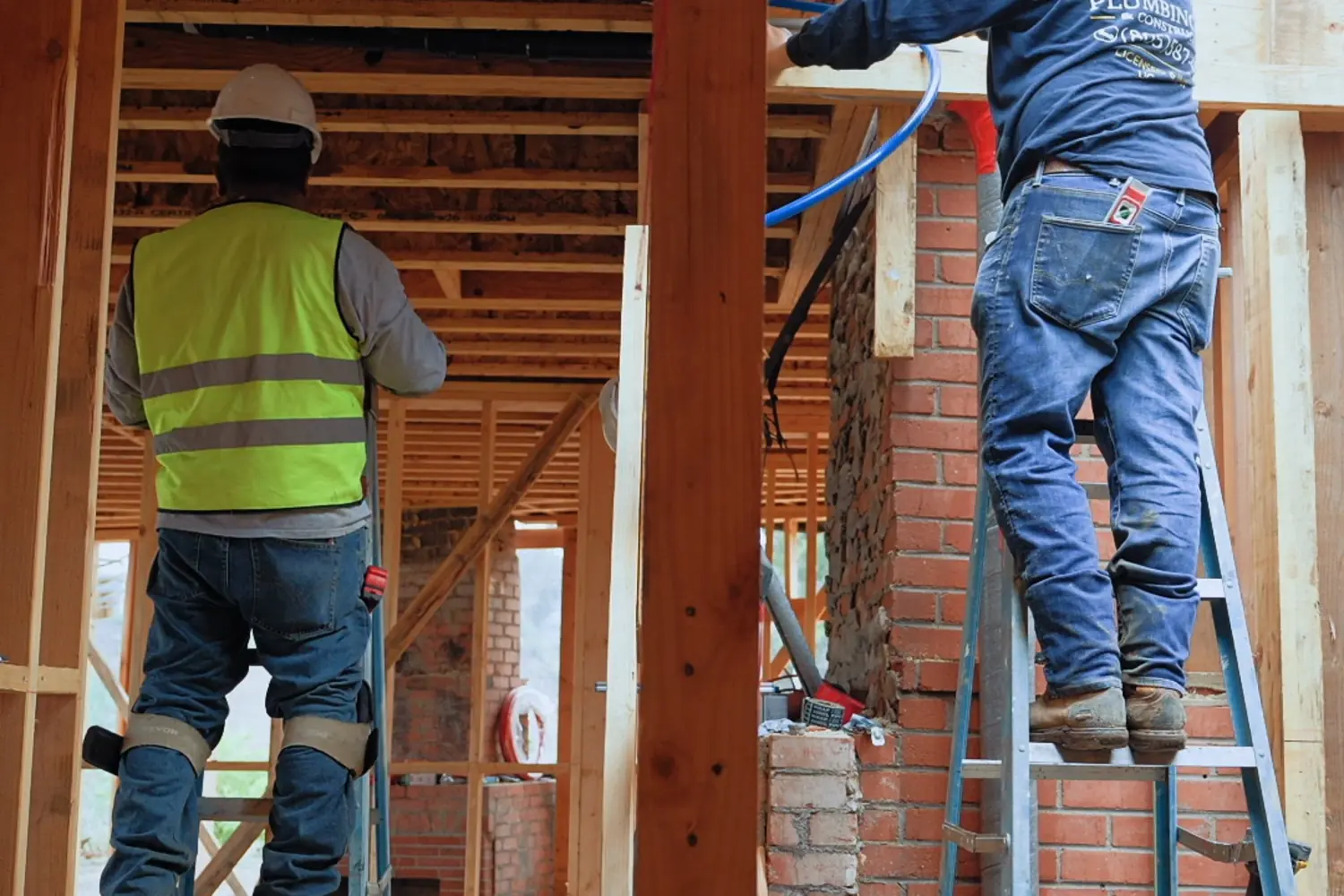 Two plumbers on ladders running pipe up a brick chase inside a framed house